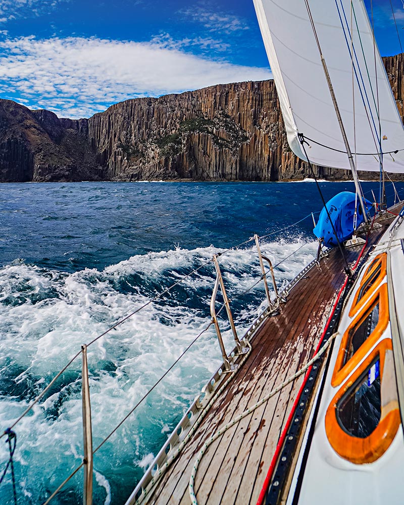 A sailboat chases the wind near Cape Raoul in Tasmania, with frothy waves crashing up against the boat.