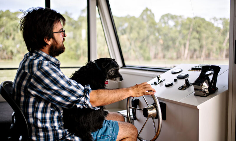 A man and his dog sit behind the wheel of a houseboat. 