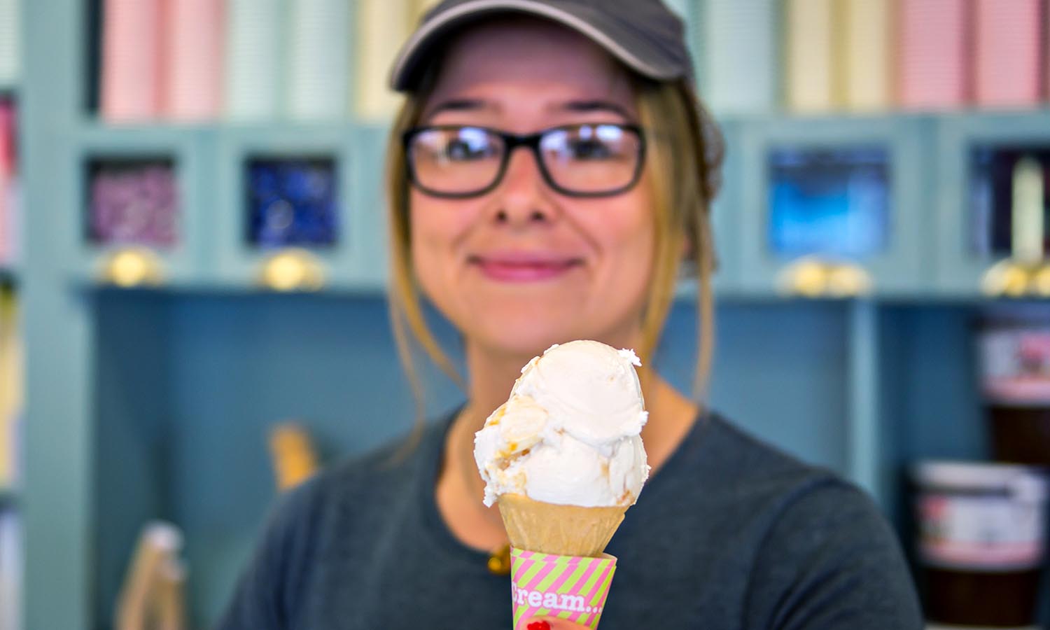 A woman smiles at the camera while holding up a cone of gelato. 