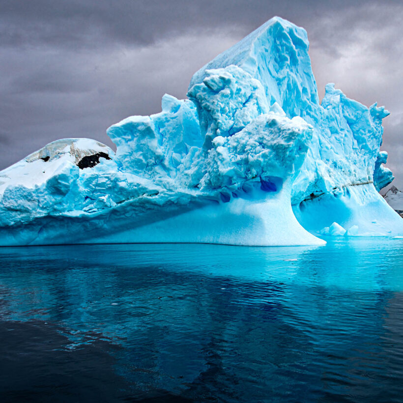 A massive blue coloured iceberg stretches away from the water's glassy surface.
