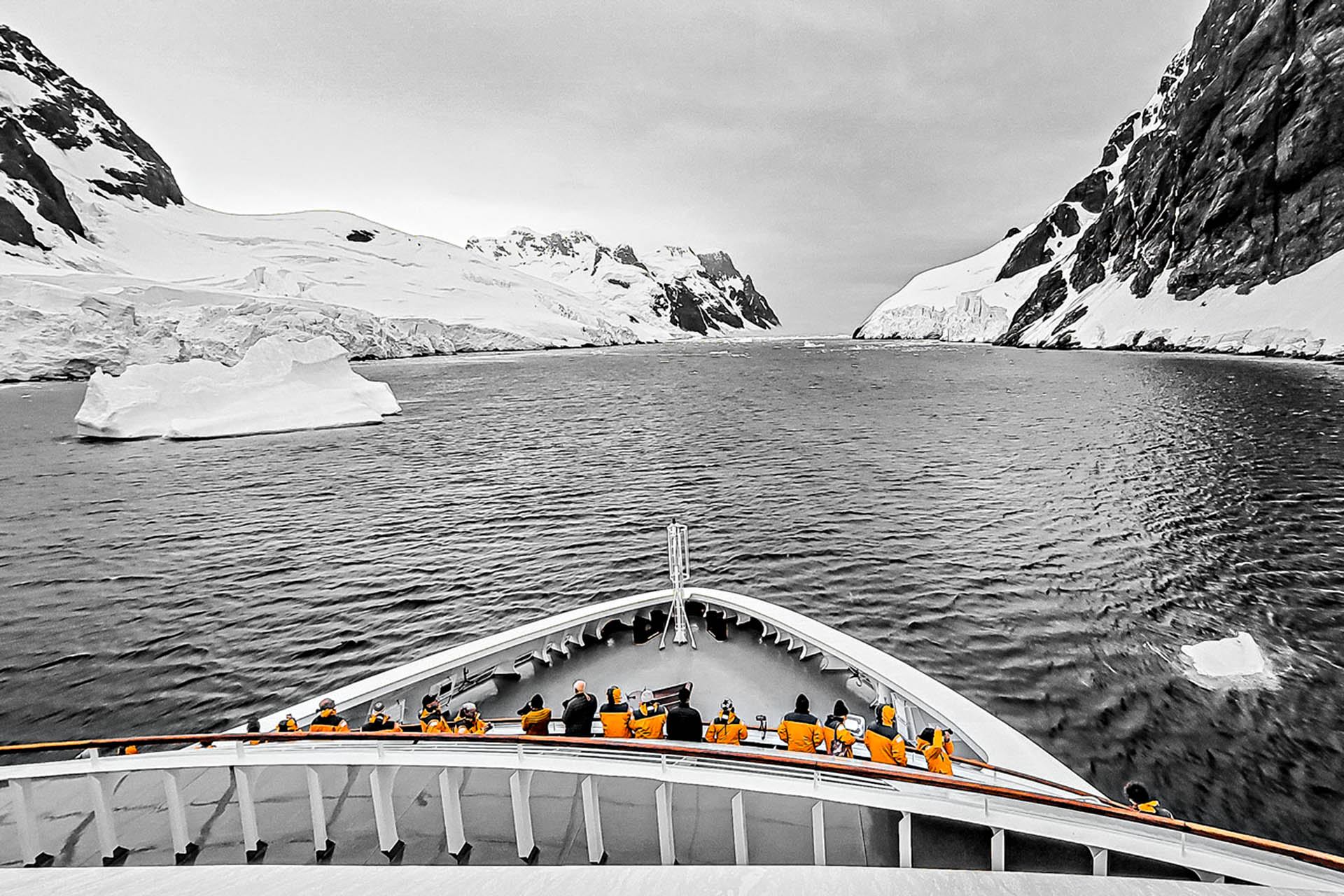 A group of people stand at the front of an expedition ship, surrounded by icebergs and icy cliff faces.
