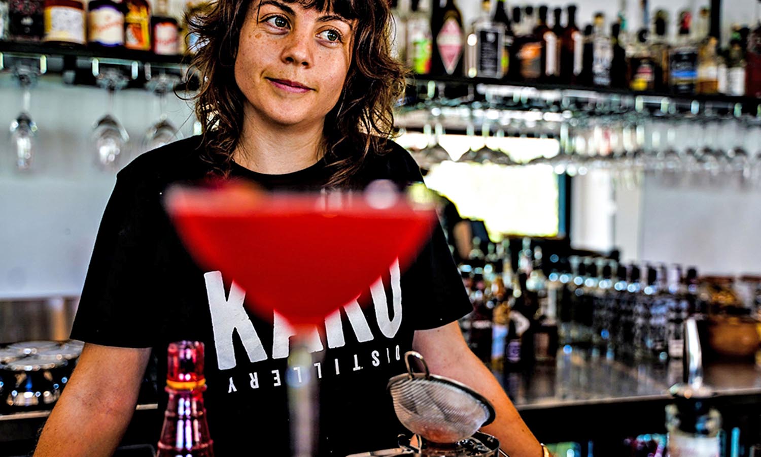 A woman stands behind a bar having just served a red coloured cocktail.