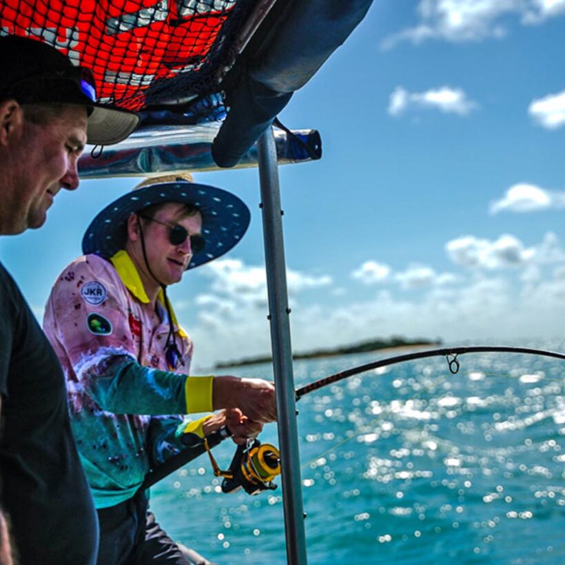 A man reels in a fish on a boat over turquoise coloured water.