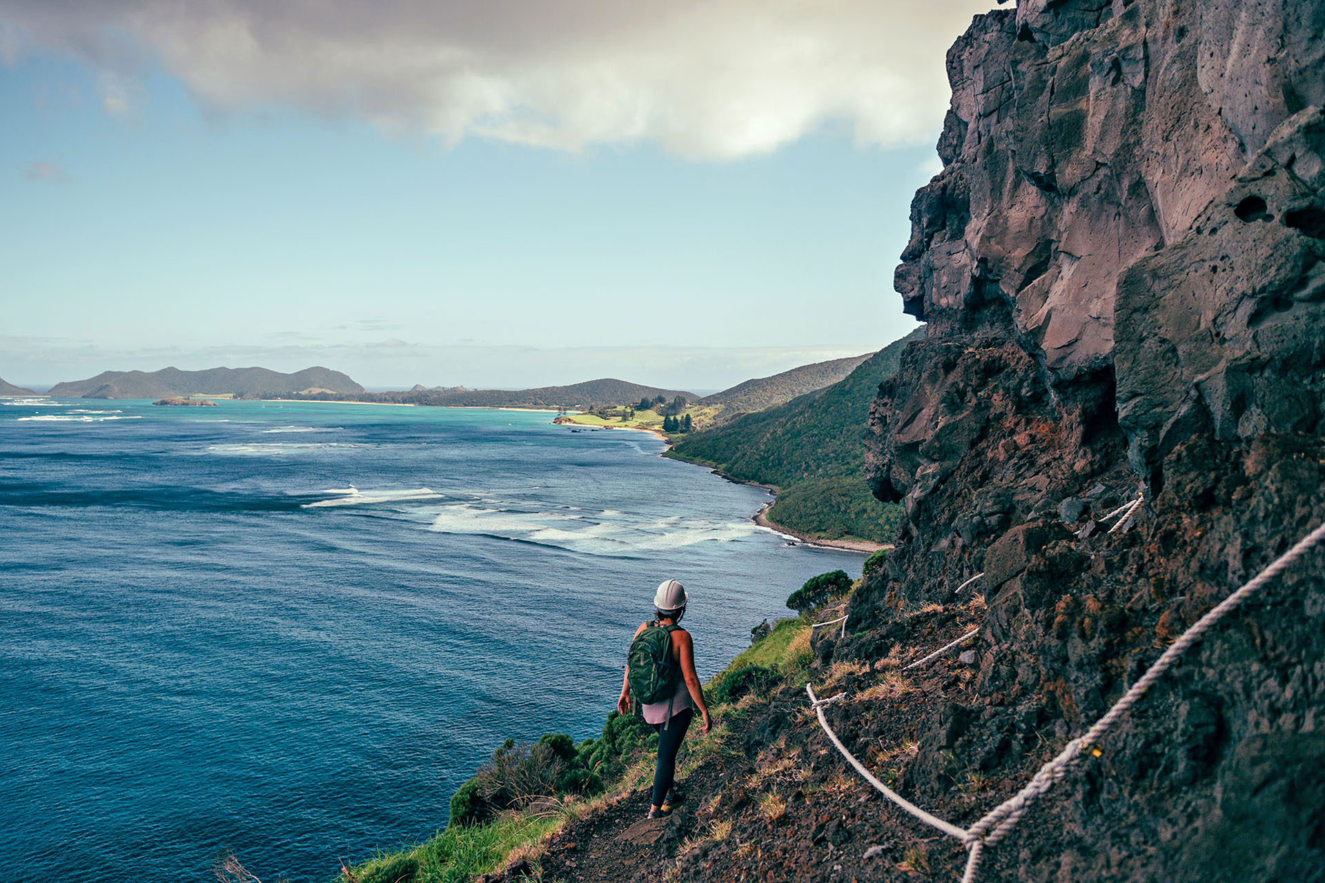 A lone person hiking around the base of a rocky outcrop with a rope to hold onto on one side and a drop to the ocean on the other.