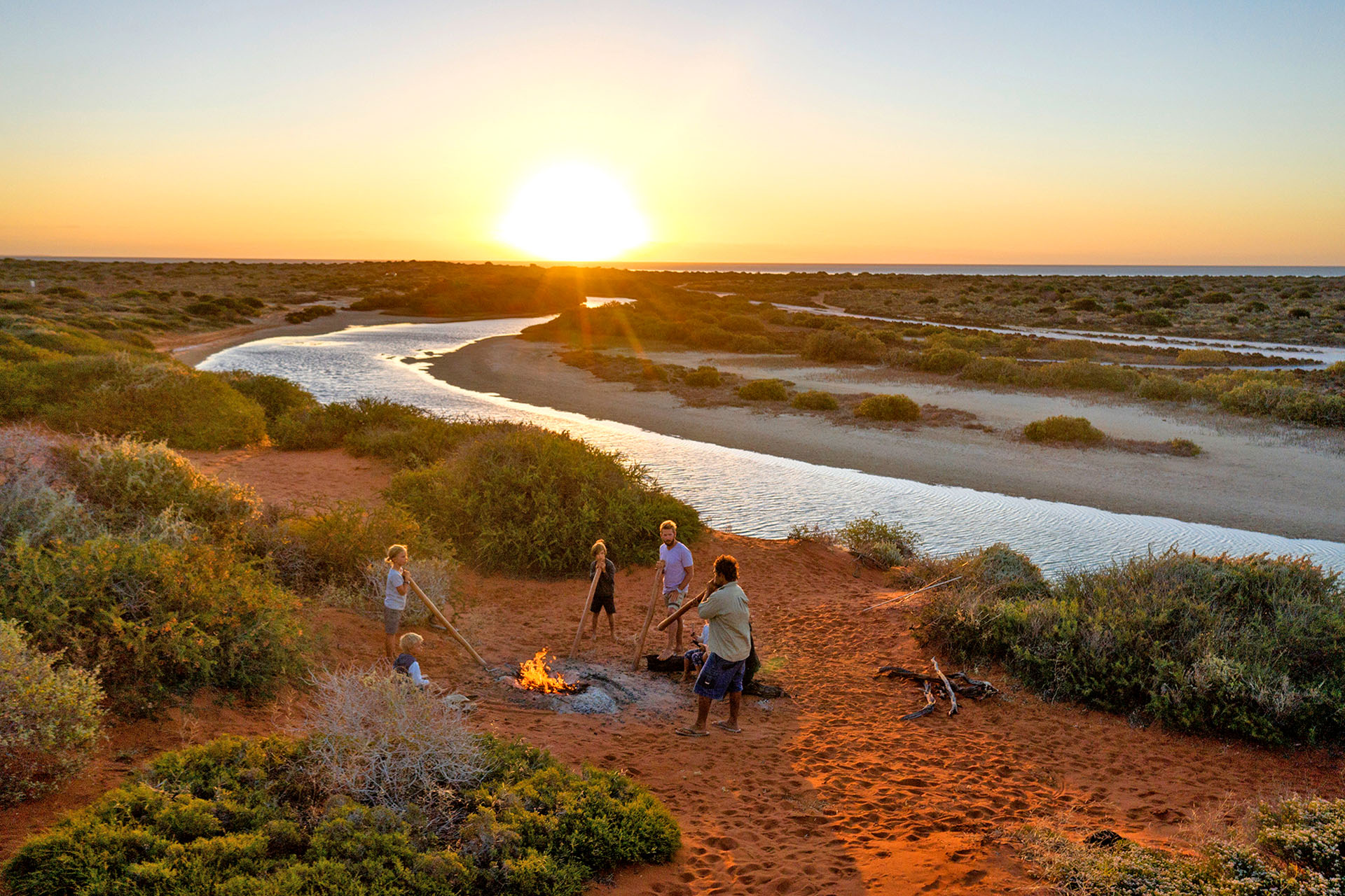 A group of people gathered around a fire pit in Western Australia, listening to an Aboriginal man playing the didgeridoo.