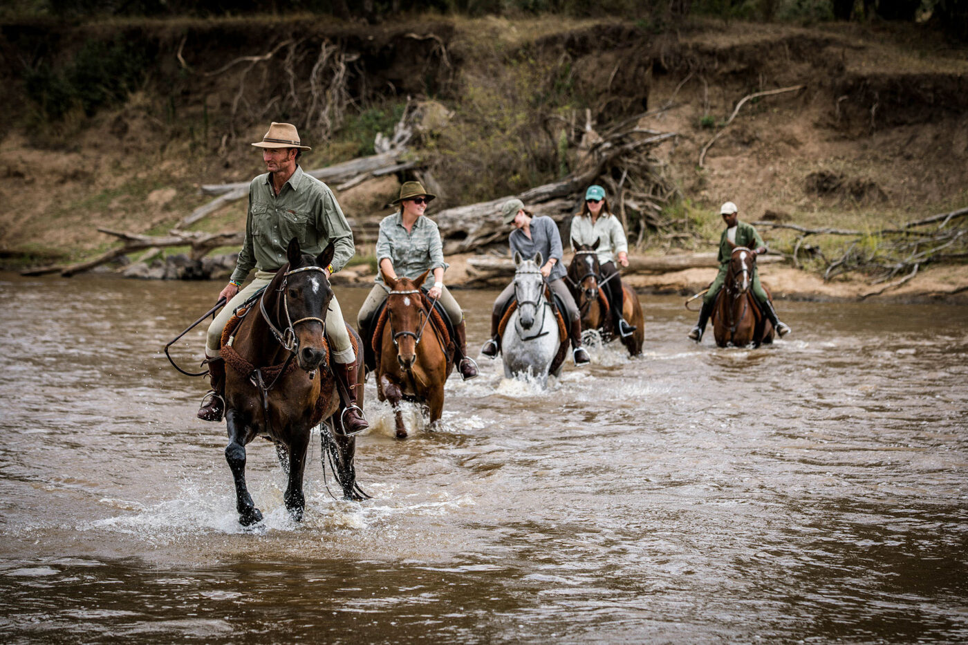 A trail of people riding horses walk through a river in Kenya.