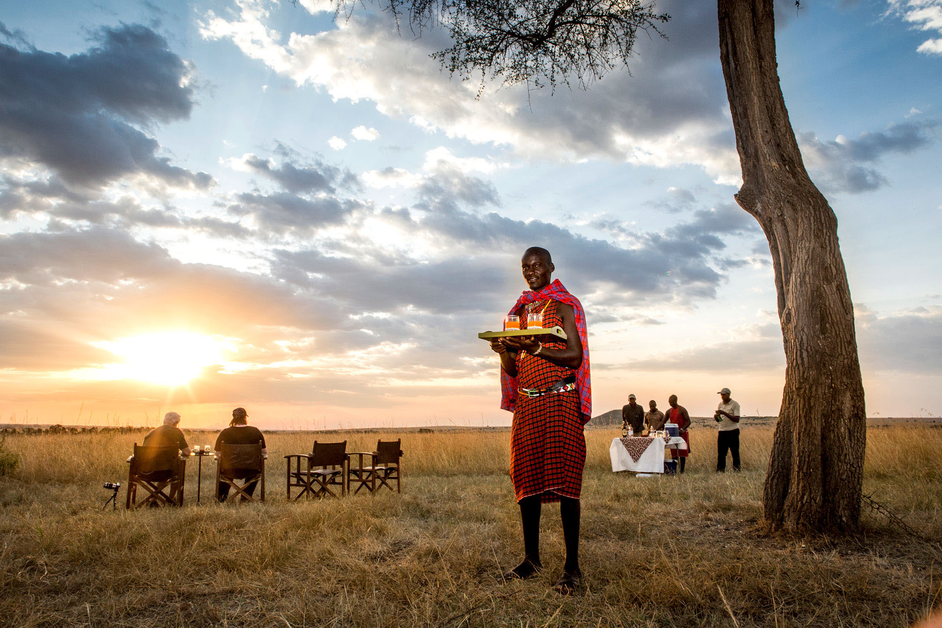 A local Kenyan man in traditional dress offering drinks as the sun sets.