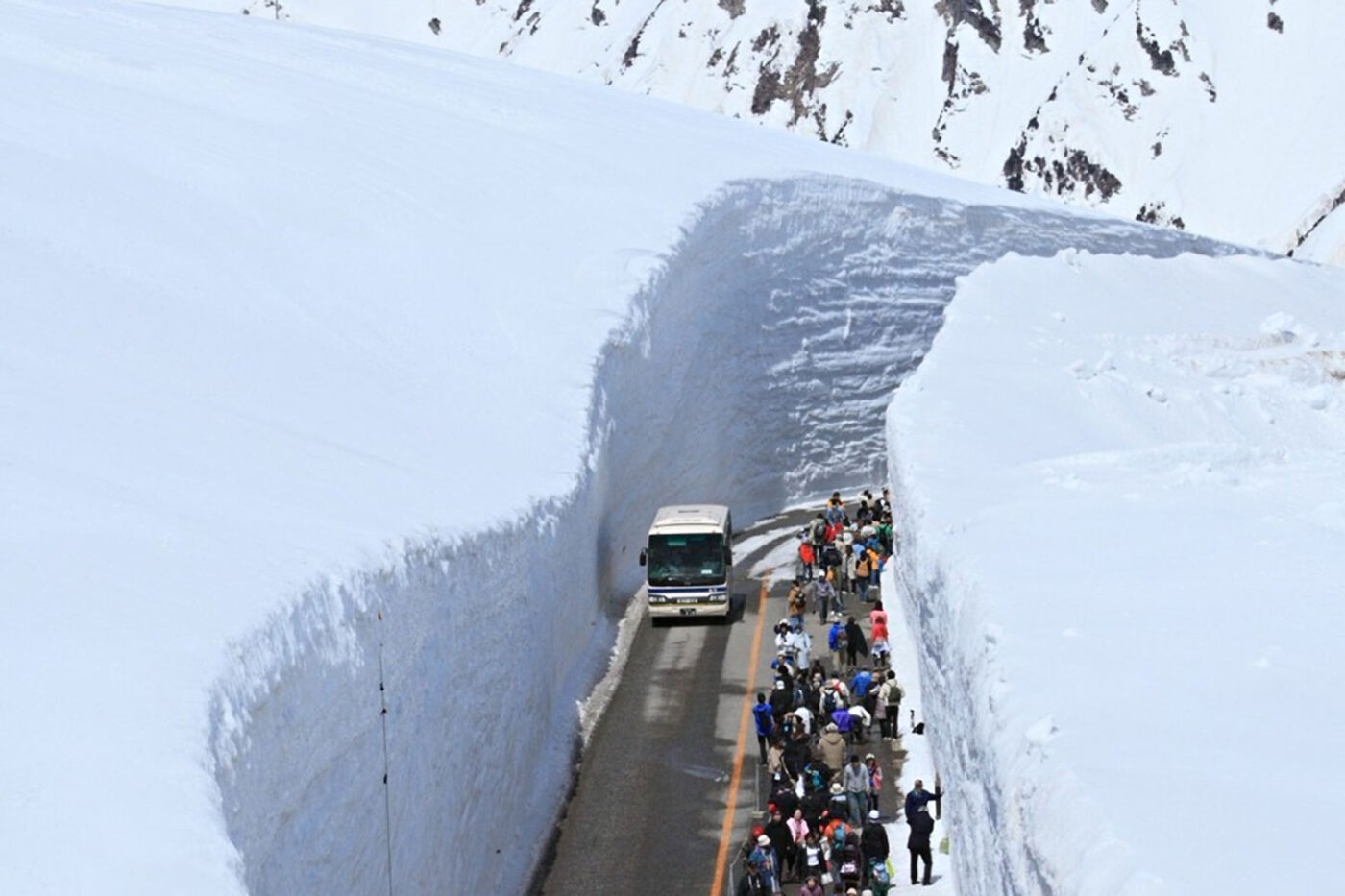 A couple of buses driving along an icy road hemmed in by more snow, as people walk beside them.