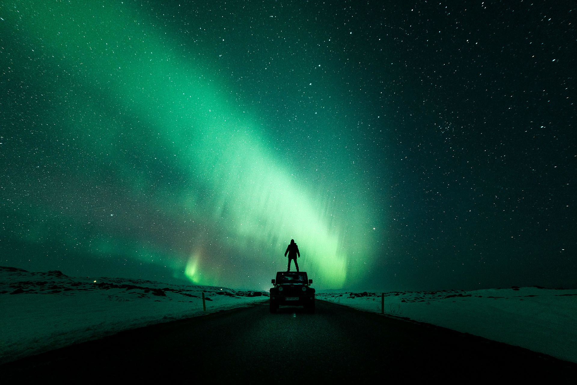 A person stands on the roof of a car underneath the colourful Northern Lights.