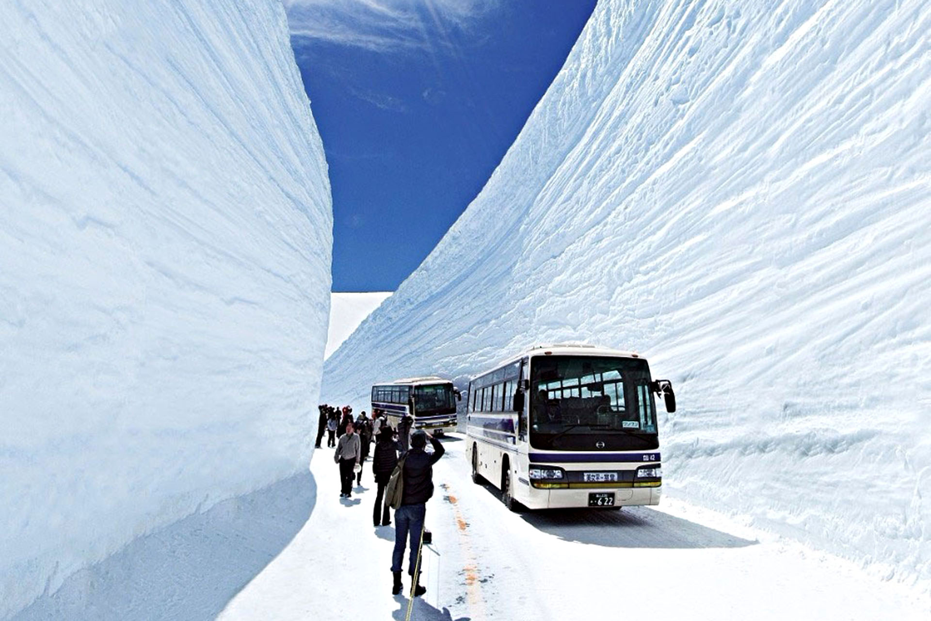 A couple of buses driving along an icy road hemmed in by more snow, as people walk beside them.
