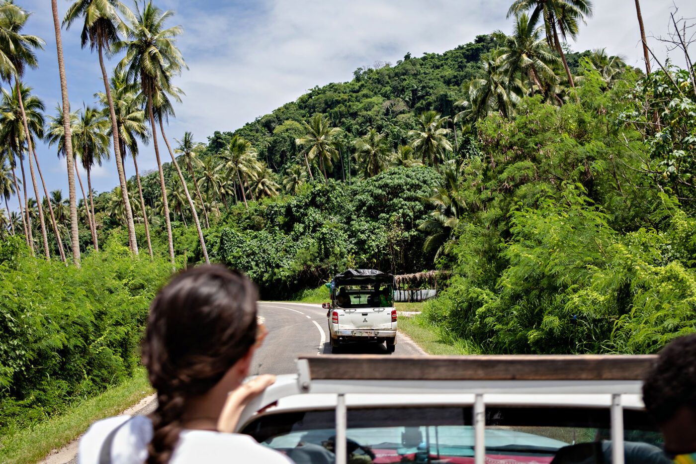 People in the back of a truck, driving along a road in Vanuatu that's lined by palm trees and lush greenery.