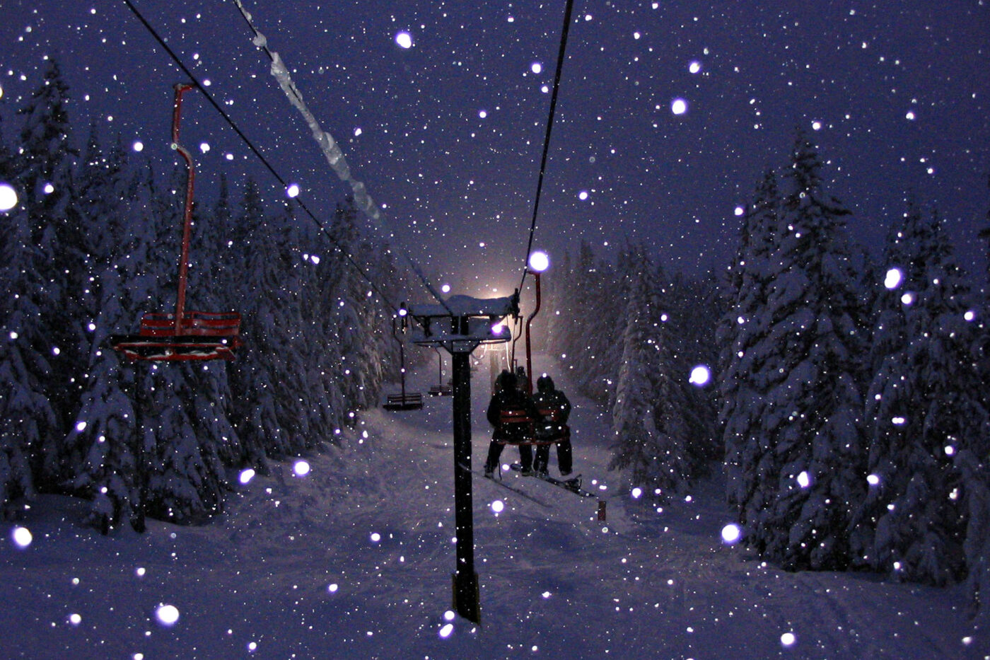 Two skiers are sitting on a chairlift while it snows at dusk.