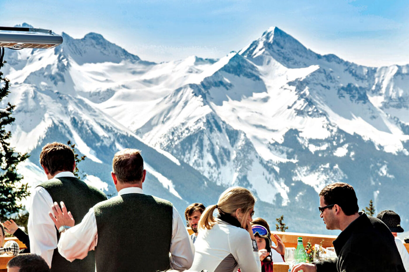 People sitting at tables outside, enjoying a meal, with snow-covered mountains in the distance.