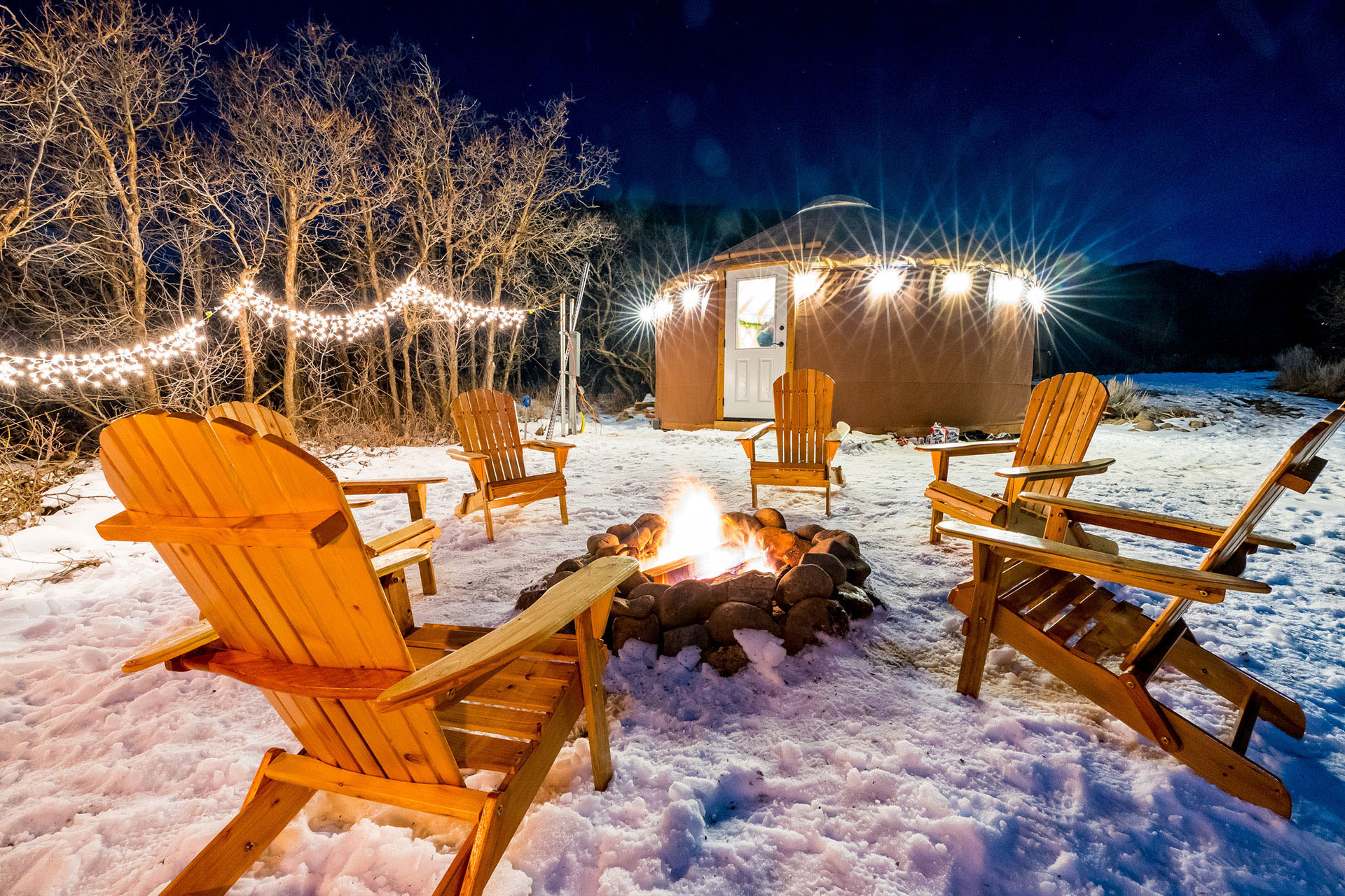 Several wooden deck chairs circle a roaring fire pit by a yurt in Midway Utah.