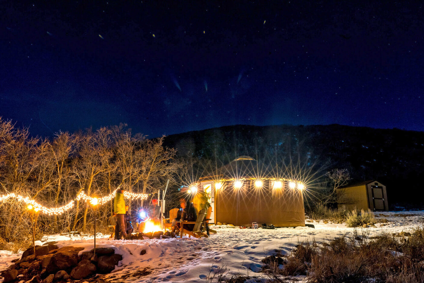 A group of people congregating around a fire pit in the snow with a traditional yurt in the background.