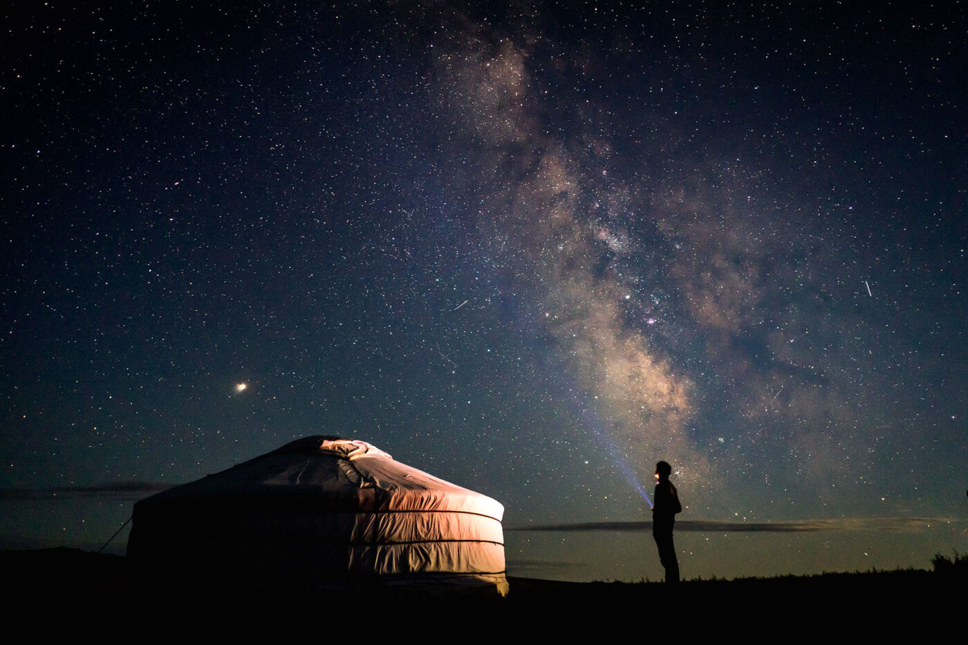 A lone man, staring up at the night sky with a flashlight, standing beside a traditional Mongolian yurt.