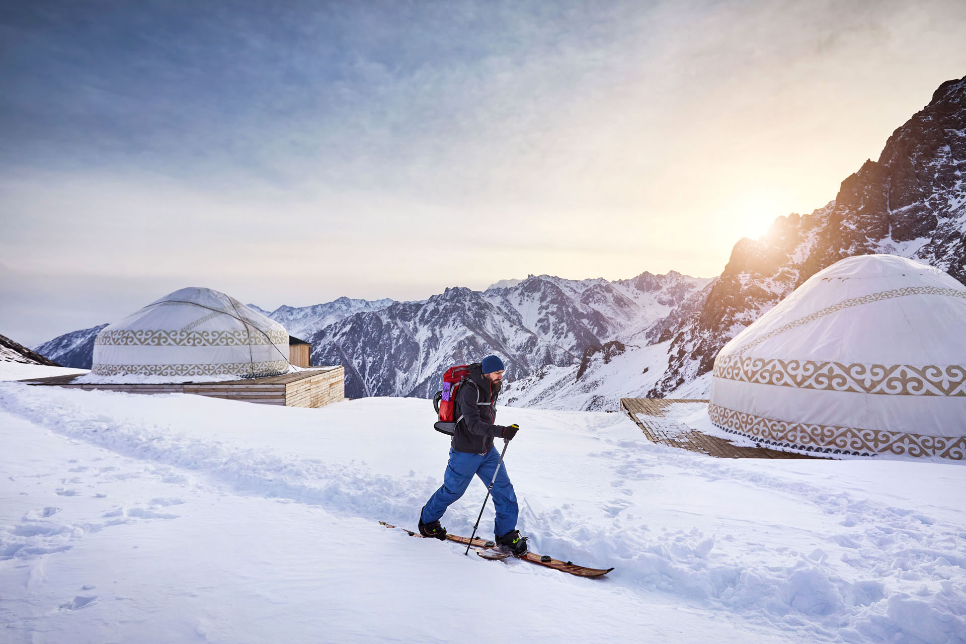A man walks through snow on a set of skis between traditional yurt accommodation among the mountains in Kyrgyzstan.
