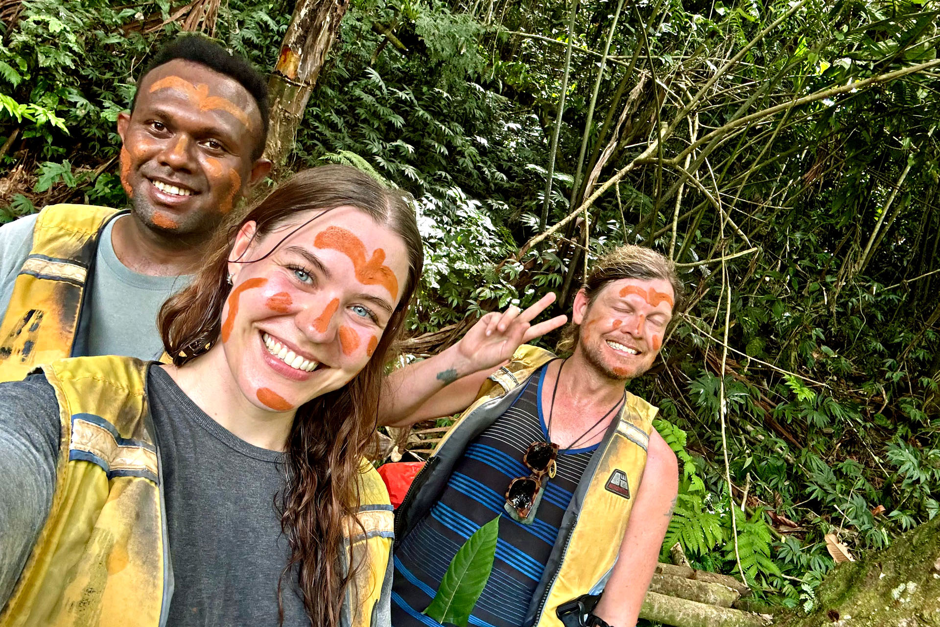 Three people with orange, muddy stripes on their faces smile and pose for the camera during the Millennium Cave trek in Vanuatu.
