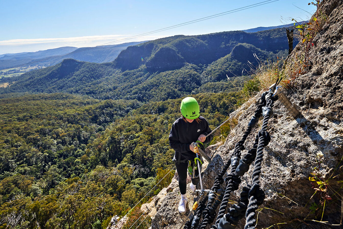 A woman climbing on the side of a cliff, doing the via ferrata at Happitat Adventure Park.