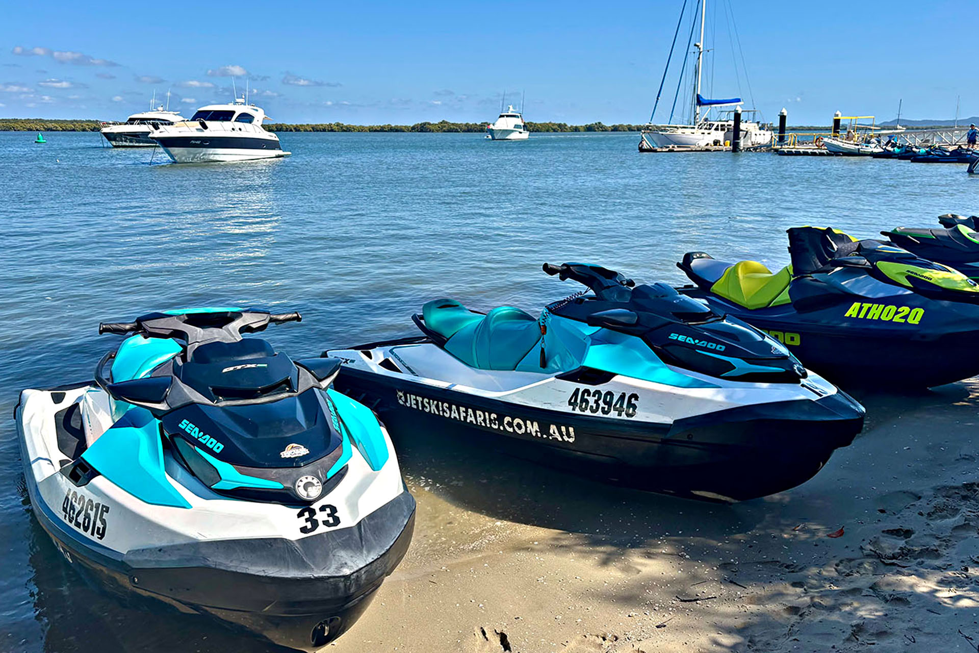 A row of jet skis lined up on the shoreline with boats bobbing on the water in the background. 