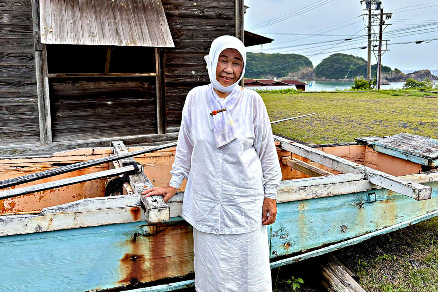 A traditional Ama diver in a all white posing by a rusty fishing boat.