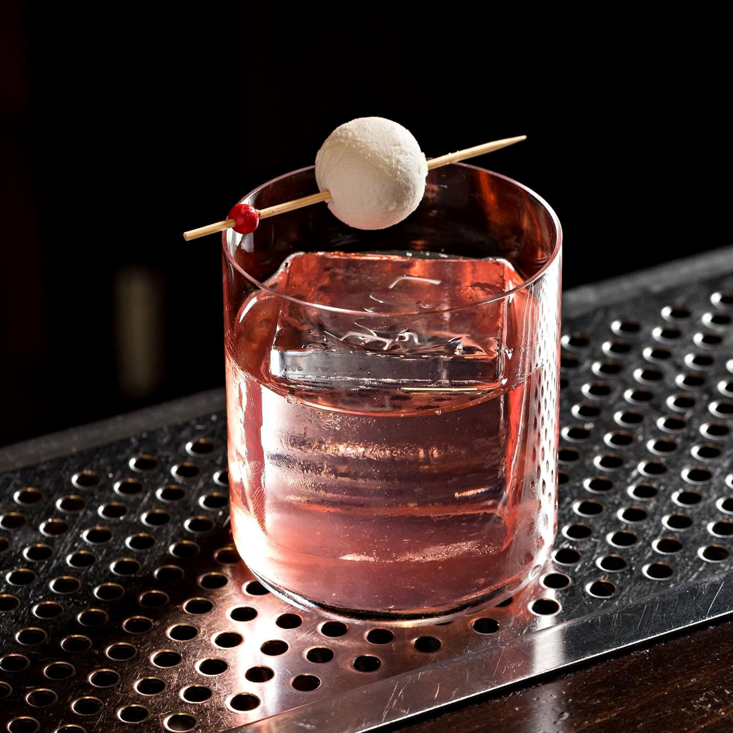 Pink signature cocktail served over a large ice cube with a white spherical garnish.