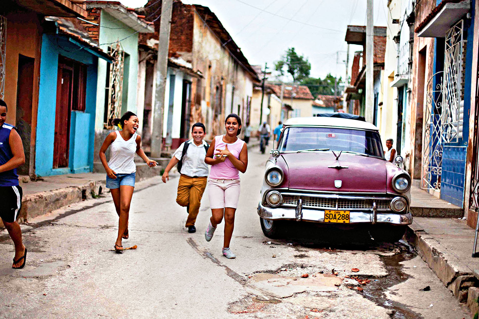 School kids run alongside an old car in a Trinidad side street.