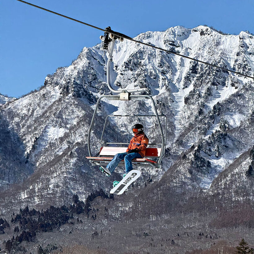Snowboarder riding a chairlift beneath the dramatic peaks of Togakushi Ski Resort in Japan.