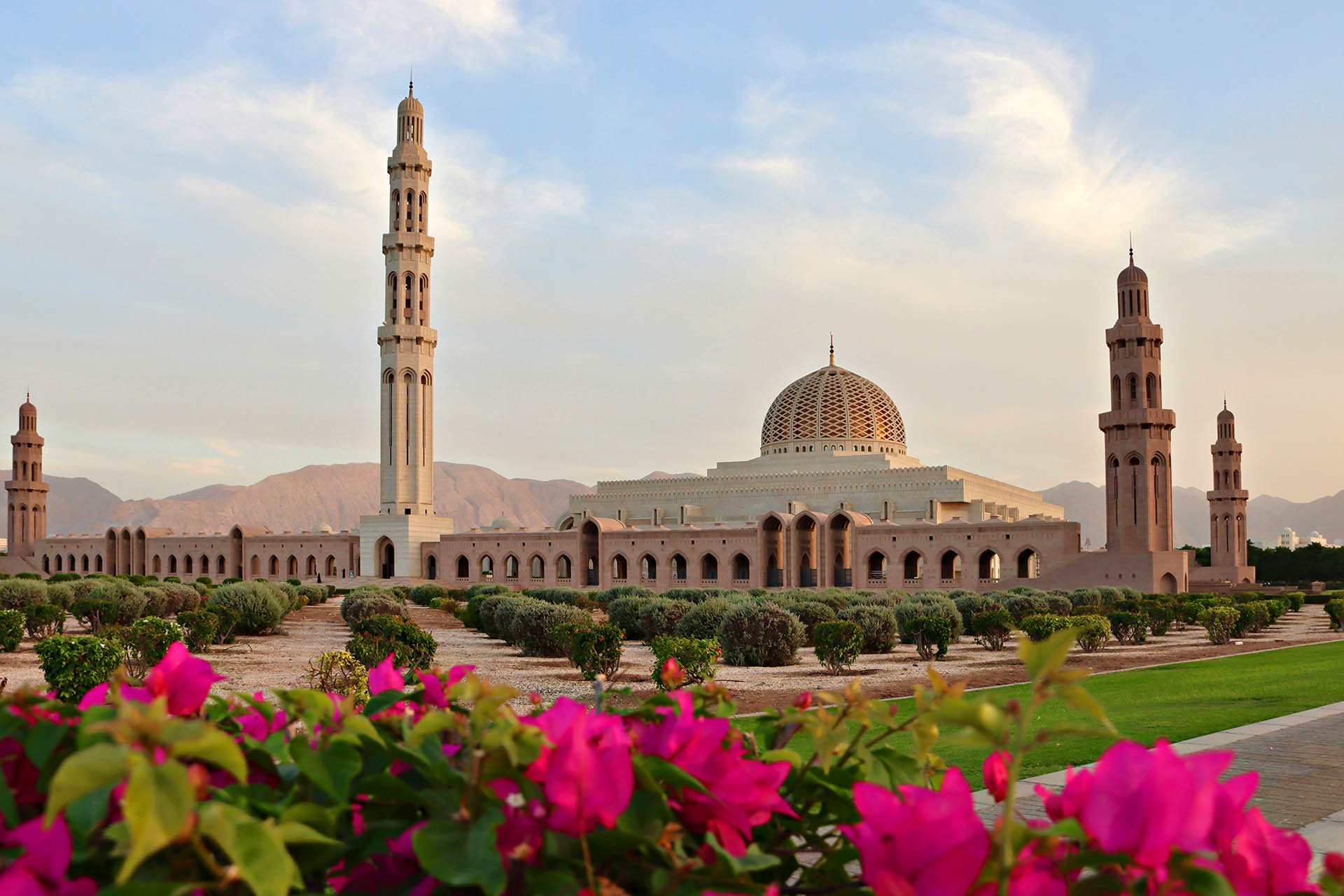 The Sultan Qaboos Grand Mosque in Muscat, Oman, framed by colourful flowers and the Hajar Mountains.
