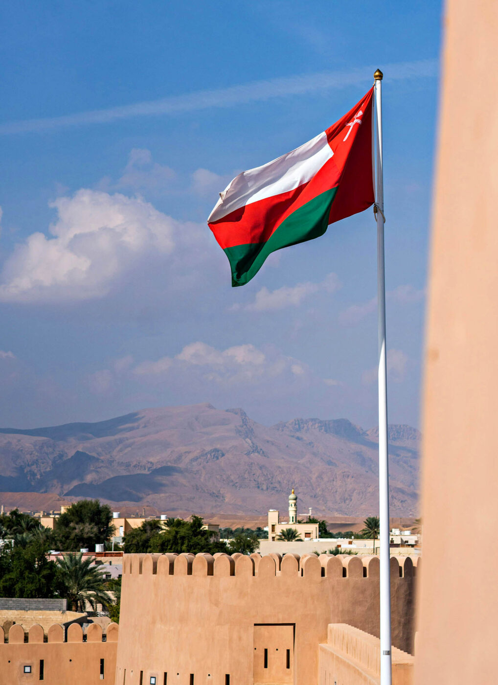 The Omani flag flying above traditional architecture with the Hajar Mountains rising behind Muscat, Oman.