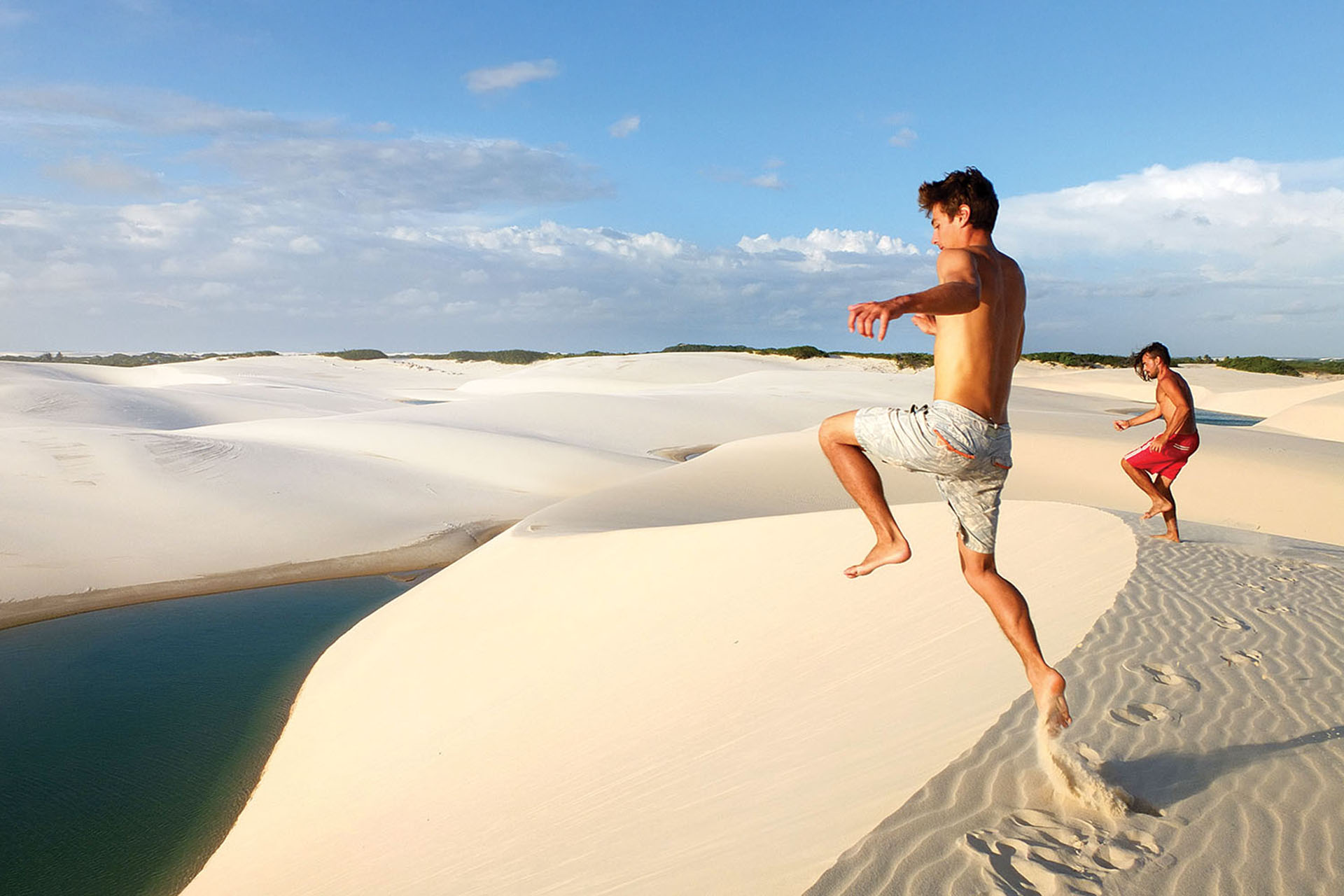 Two travellers running down white sand dunes beside a lagoon in Lençóis Maranhenses, Brazil.