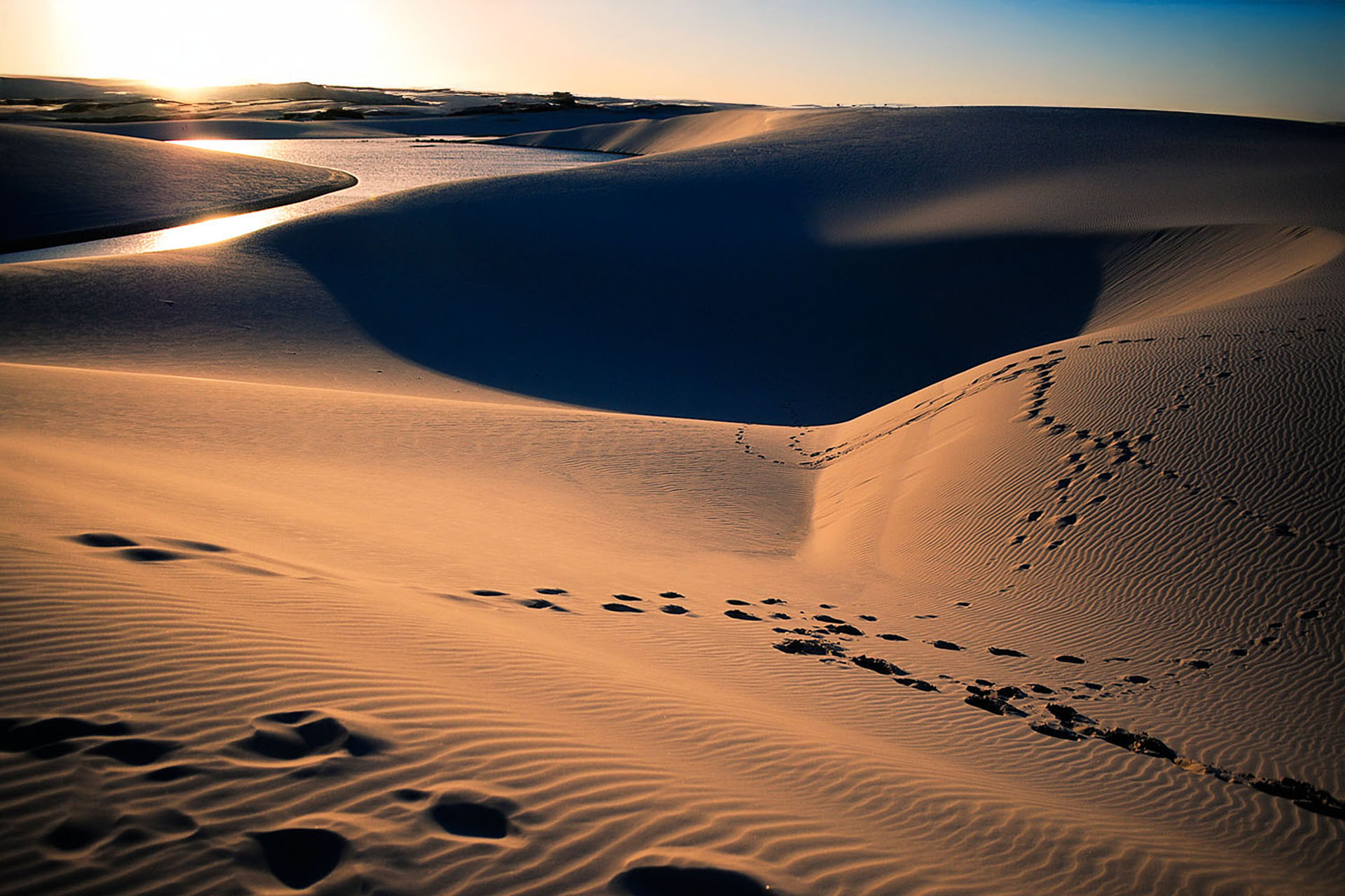 Footprints winding across sand dunes beside a lagoon at sunset in Lençóis Maranhenses.
