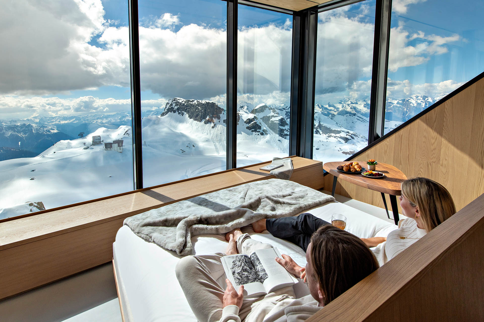 Guests relaxing in a Cabane Tortin bedroom with floor-to-ceiling views across the snow-covered Swiss Alps.