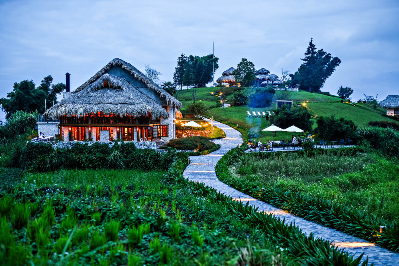 Topas Ecolodge Sapa at dusk with stone paths, thatched-roof buildings and gardens in the mountains of northern Vietnam.