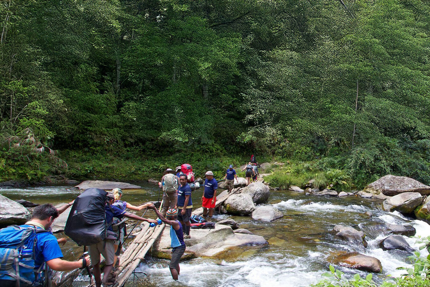 A group of people walking over small planks of wood, trying to cross a river.