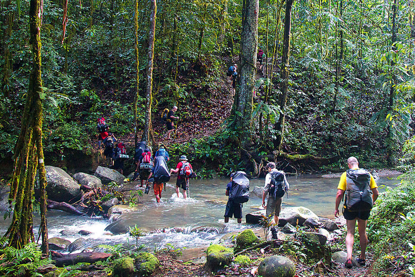 A big group of people crossing through a river, getting wet up to their knees.
