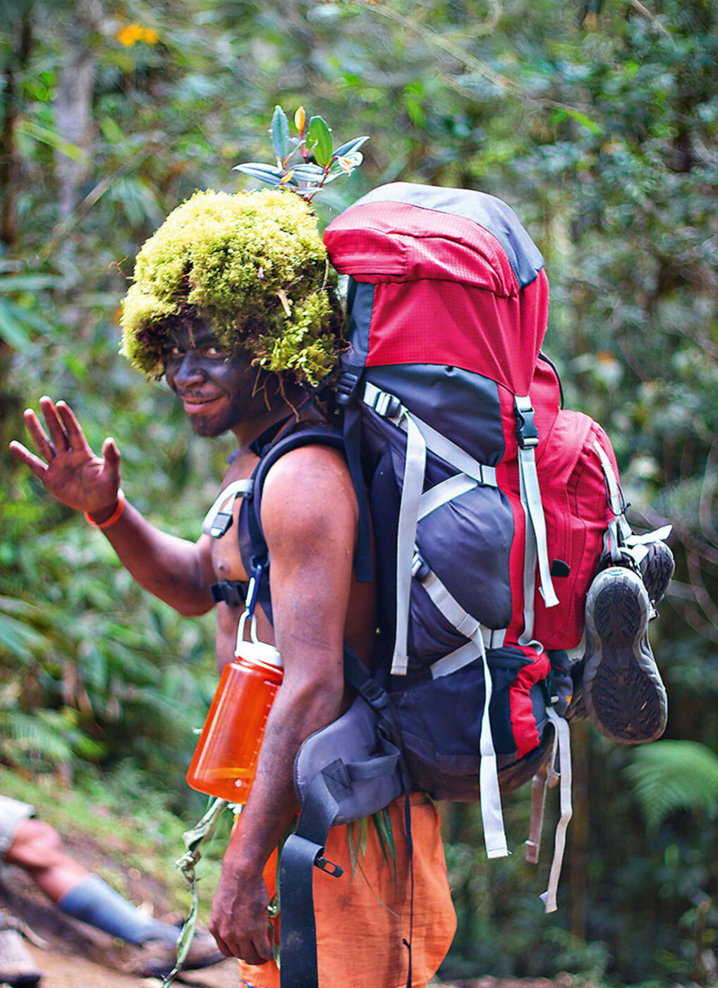 A person with a big hiking pack on their back and a moss hat on their head, waving to the camera.