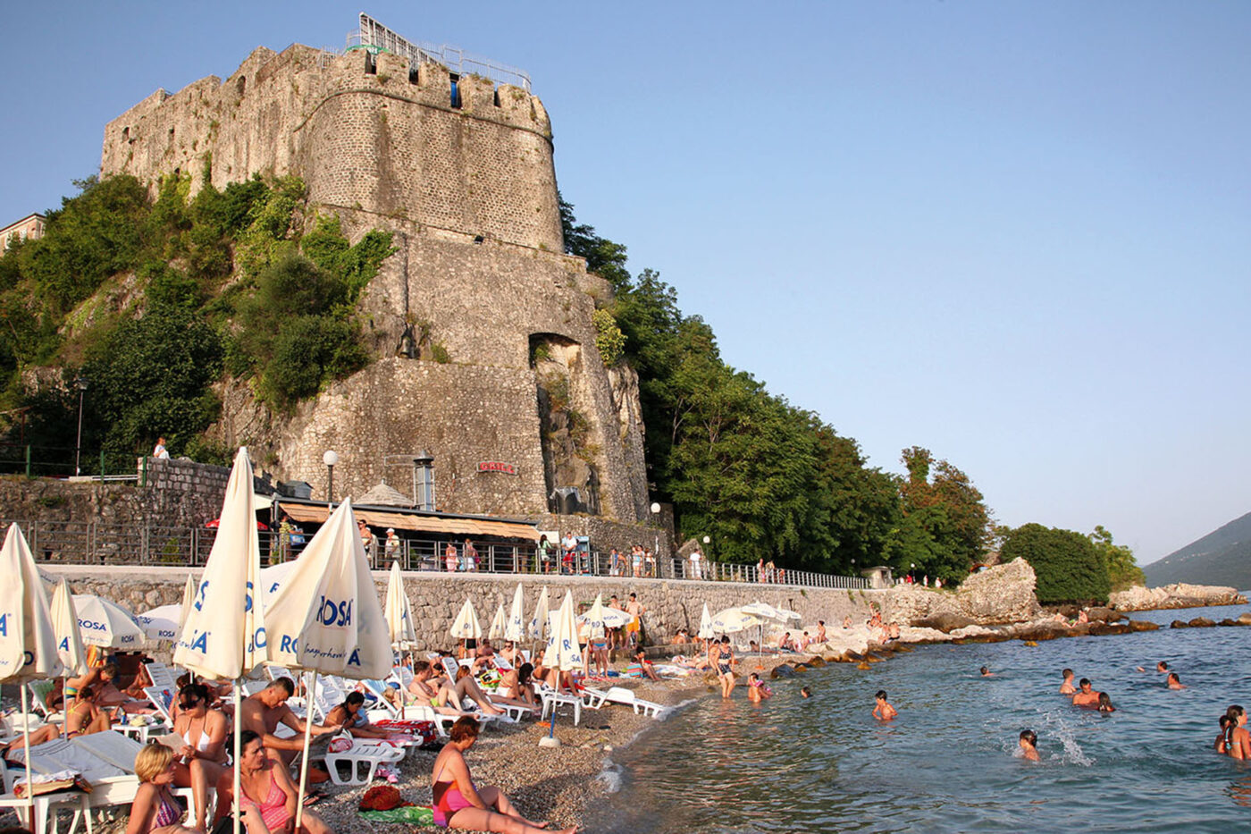 A stone beach full of people. There's a big stone building overlooking the sea.