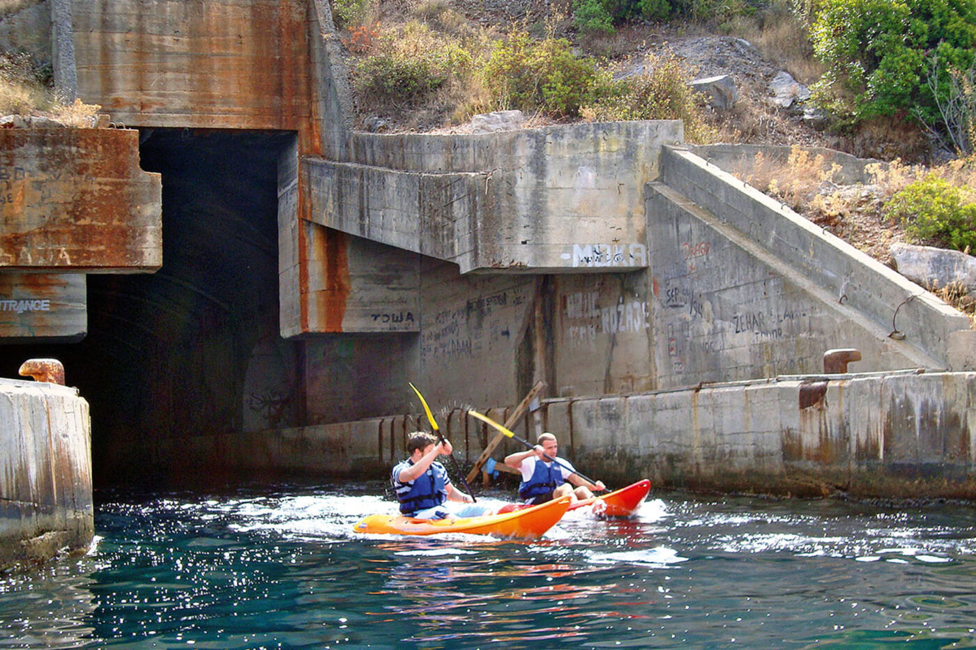 Two people in orange kayakers emerging from a grey submarine pen.