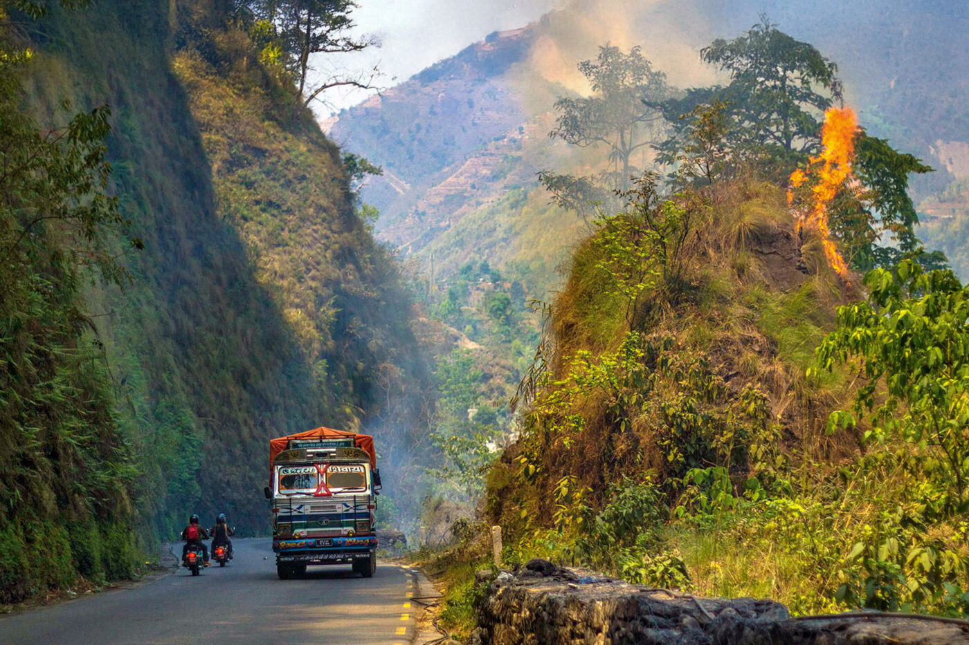 A long road through the mountain with a truck driving towards the camera, and two motorcyclists driving away from it. Next to this, a hill of greenery with a tree that's on fire.