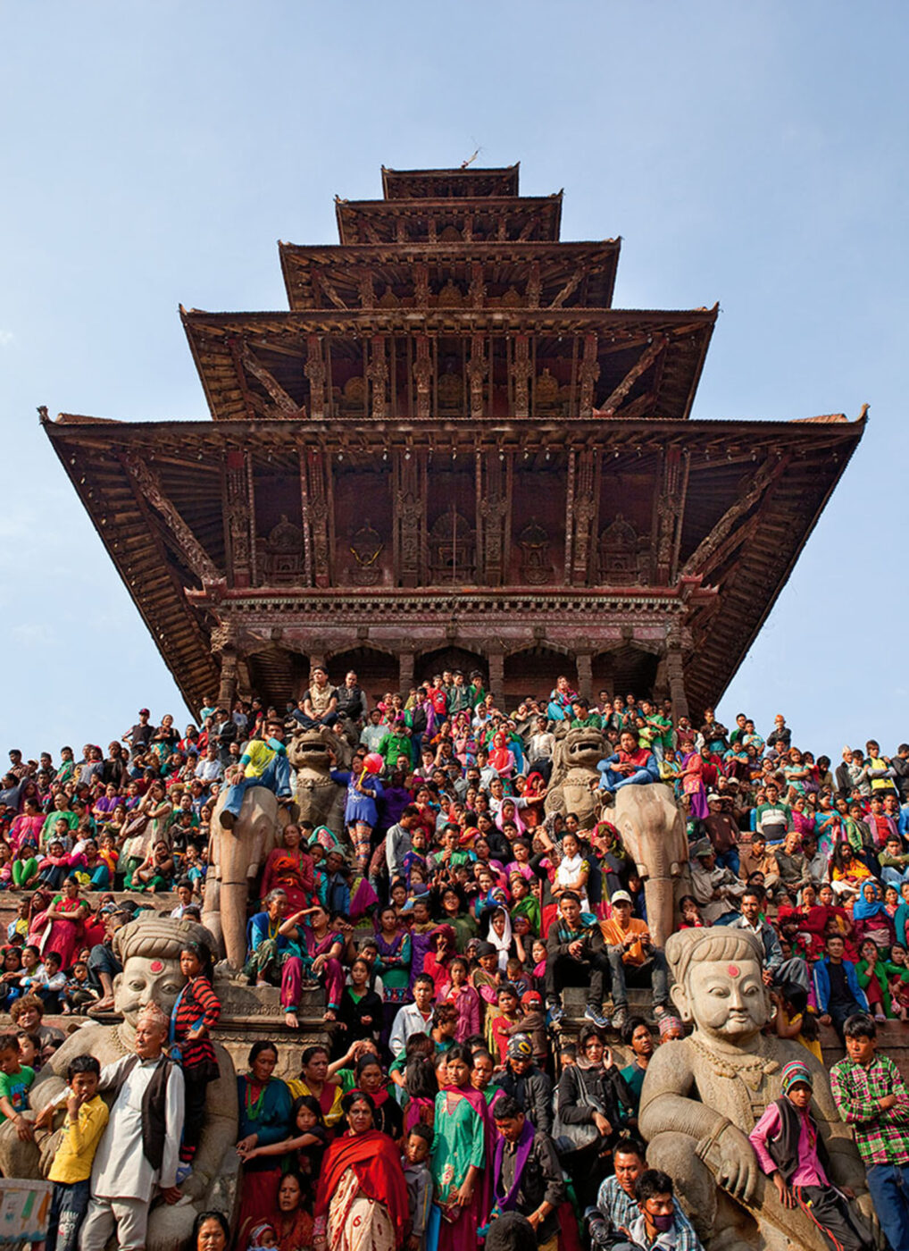 A big group of people sitting on the stairs outside a temple. With six big statues scattered around them.