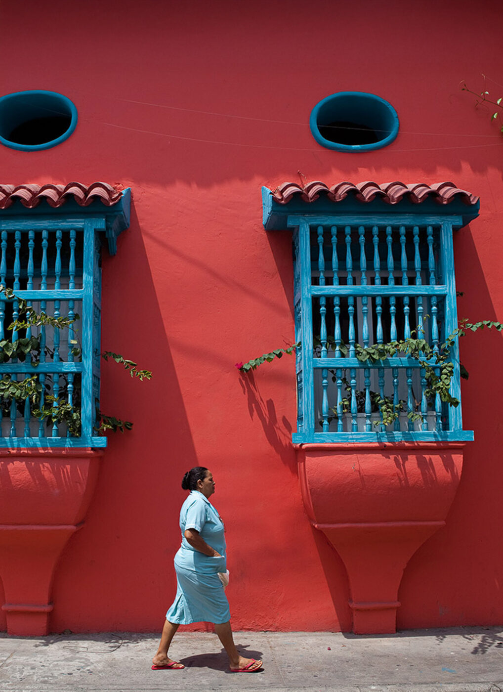 A woman walking past a bright red painted building with bright blue fences on the windows.