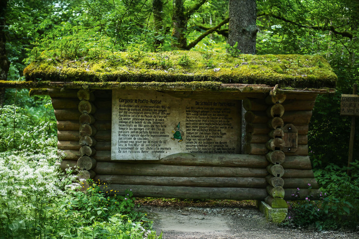 A sign on a log cabin with text in two different languages that tells the story of a green fairy.