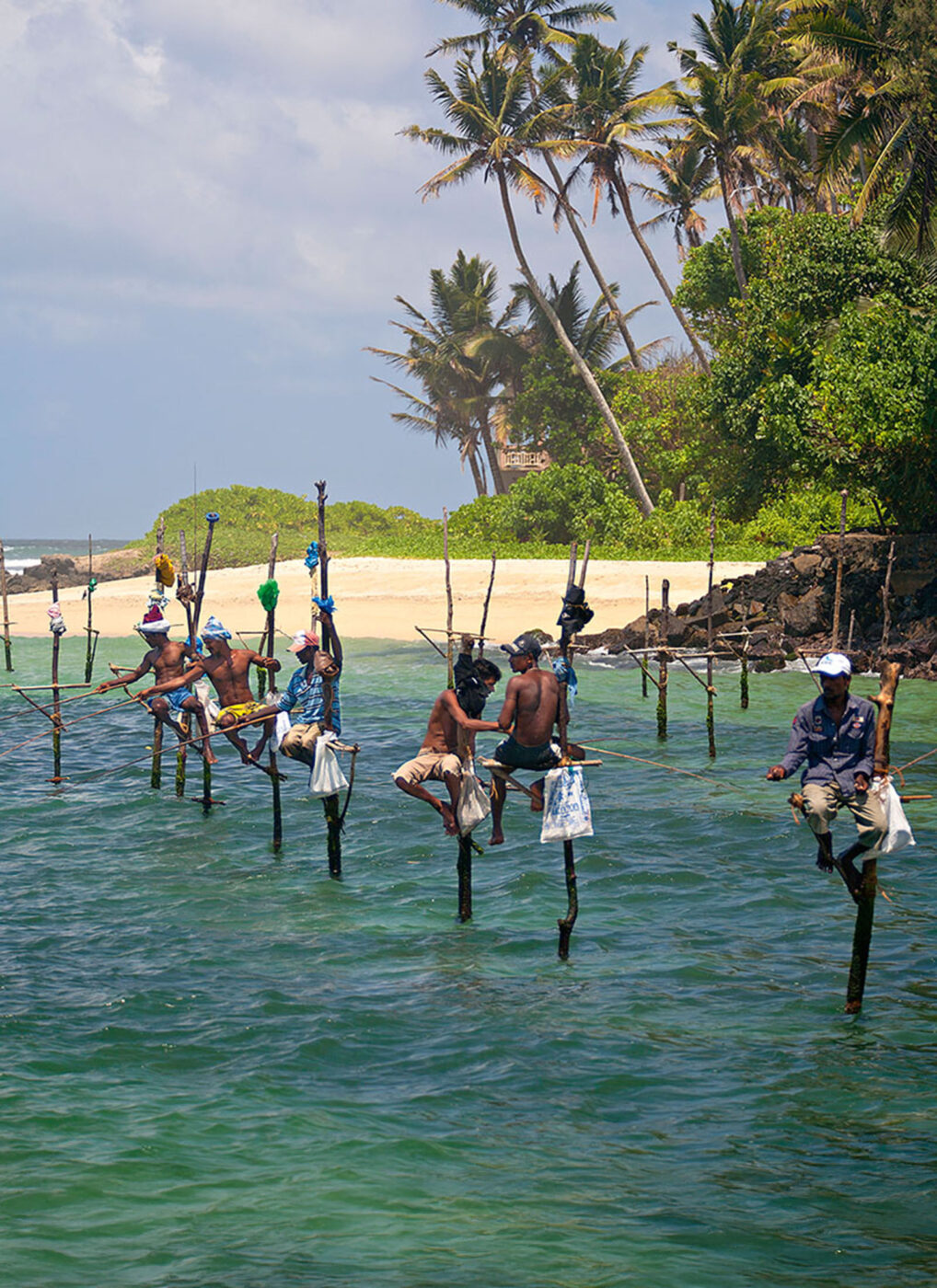 A group of 7 fisherman hanging on big stilts in the water with their fishing rods in the water.