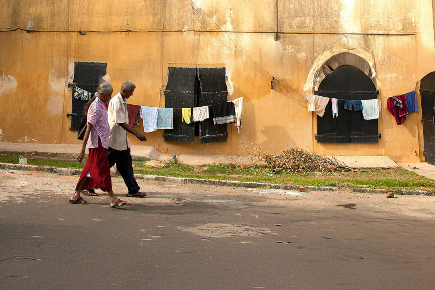 Two people walk down a road next to a brown building with clotheslines hung onto the side of it.