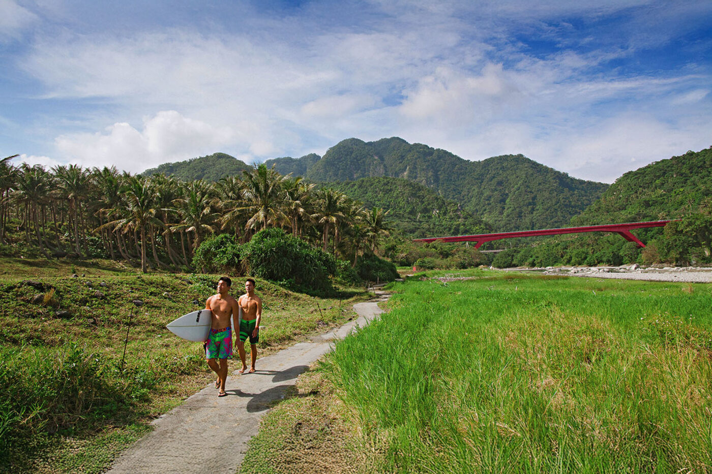 Two men holding surfboards walking the path to the beach. Behind them, a big red bridge.