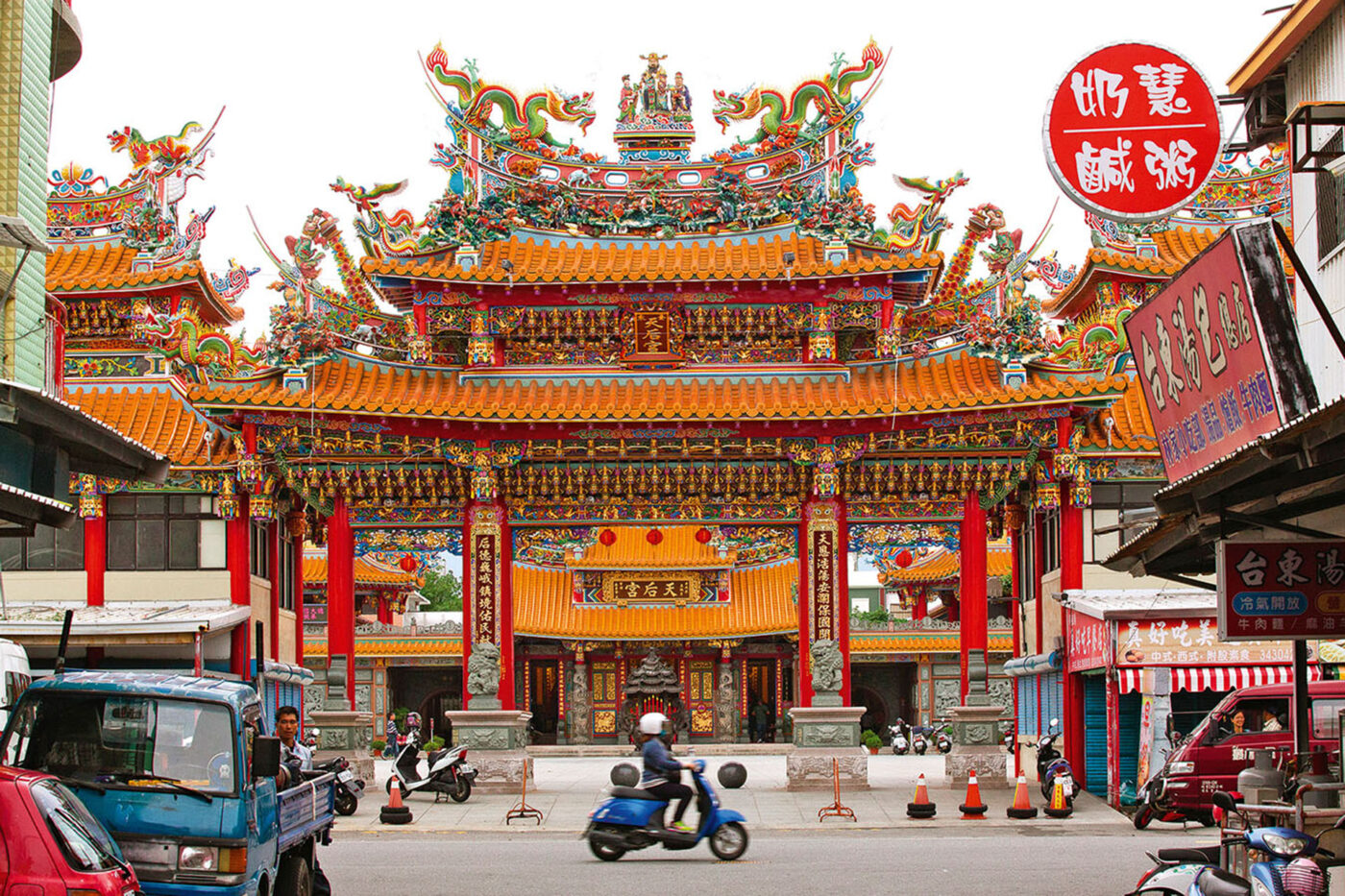 A very detailed, orange sea temple in the middle of a busy street. People on motorbikes and cars are riding past it.