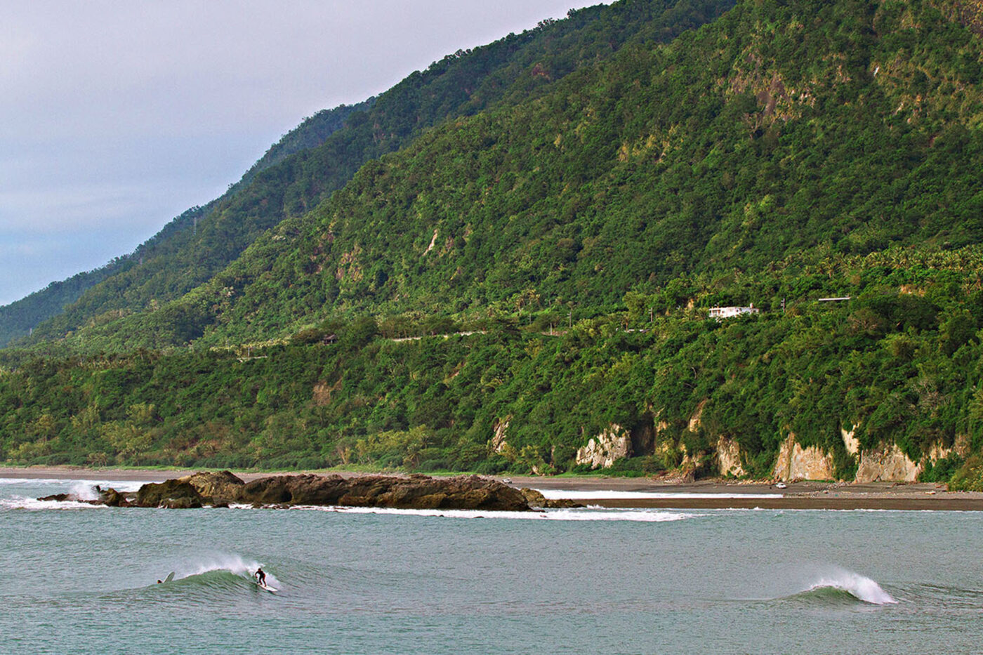 A landscape shot of the sea, rocks and the greenery around the highway up the hill. In the water, a surfer is riding waves.
