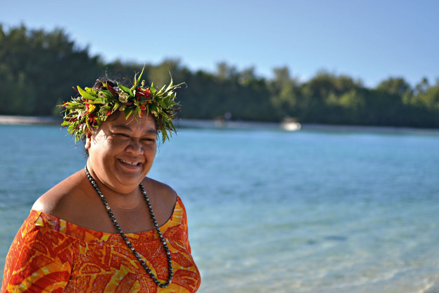 A woman wearing a headpiece of leaves and honeysuckle, a black pearl necklace and an orange patterned top, smiling at the camera. In the background, a light blue ocean and greenery.