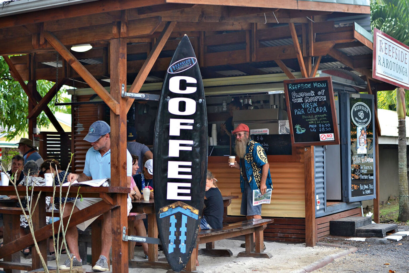 A little café with a big 'coffee' sign out the front. The tables are filled with people drinking their coffees and chatting. A man stands next to the sign with a coffee, a cigarette and a newspaper.